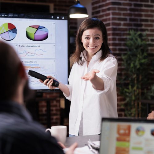 Smiling business employee standing in front of tv screen with sales statistics talking with man with laptop sitting in late night meeting. Woman presenting pie charts and analytics to mixed team.